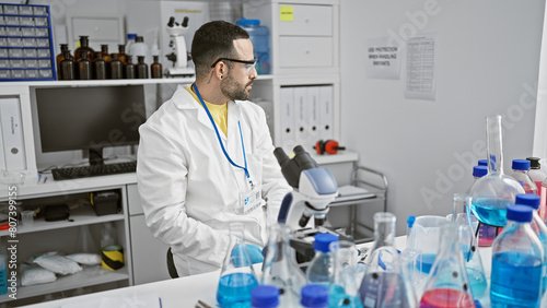 Hispanic man in a laboratory wearing a lab coat with glasses  observing scientific equipment indoors.