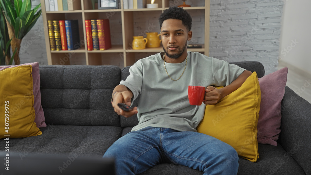Relaxed african american man holding remote and coffee mug on cozy home couch in a living room.