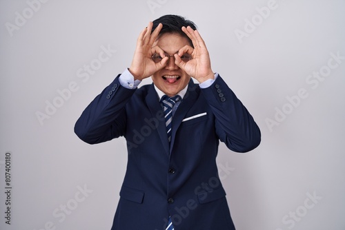 Young asian man wearing business suit and tie doing ok gesture like binoculars sticking tongue out, eyes looking through fingers. crazy expression.