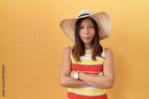 Middle age chinese woman wearing summer hat over yellow background skeptic and nervous, disapproving expression on face with crossed arms. negative person.