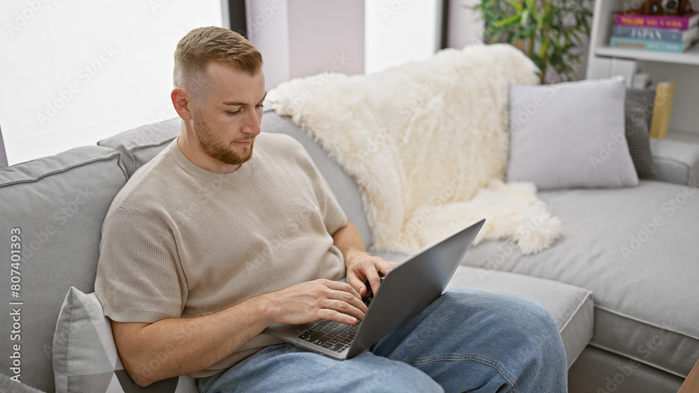 A focused young man with a beard comfortably using a laptop indoors, in a cozy modern living room.