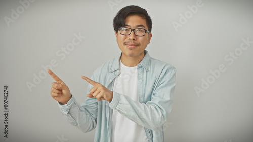 Obraz na plátně A young asian man in casual attire confidently points to the side on a white isolated background