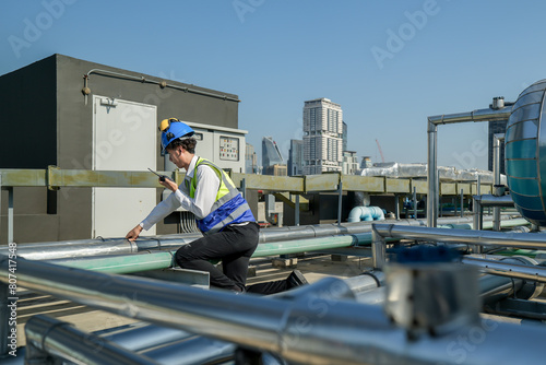 An engineer attentively checks pipework on a rooftop against a cityscape, using a tablet for data analysis in urban maintenance.