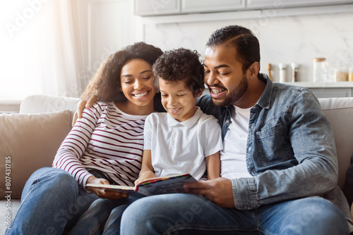 Black family consisting of parents and children are seated together on a couch, engrossed in reading a book. The parents and children are focused on the pages, expressing interest and engagement.
