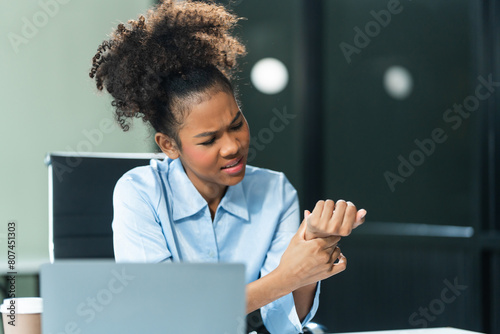 A young African American woman in a blue formal shirt with afro brown hair battles office syndrome, including wrist pain, repetitive stress, arthritis, and carpal tunnel syndrome in a modern office.