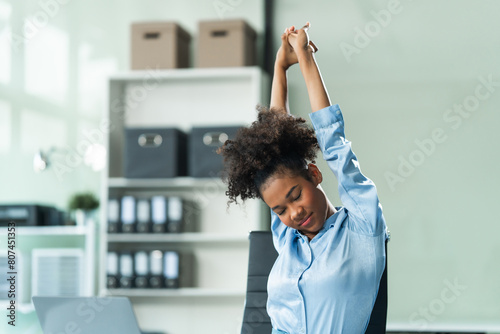 A young African American woman in a blue formal shirt with afro brown hair experiences office syndrome, tiredness, and pain from prolonged sitting in a modern office environment. photo