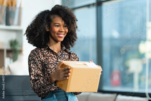 A young African American woman with afro brown hair works in a modern office, managing her online clothing store and live stream platform, sme box packages. © NanSan