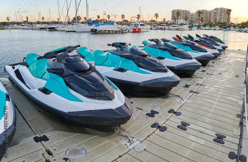 Fototapeta Naklejka Na Ścianę i Meble -  Rental jet skis parked in a row on a floating dock in a marina at sunset. Concept of water sports and activities to enjoy in the sea.