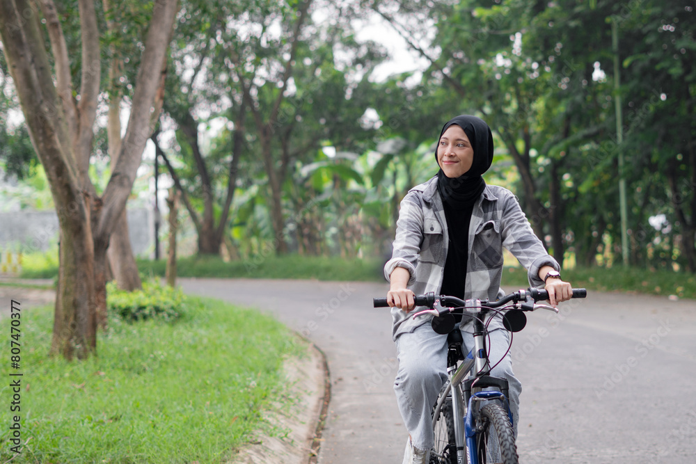 Young cheerful hijab woman, wearing casual flannel riding bicycle bike on sidewalk in city enjoying being outdoors, look aside. People active urban healthy lifestyle cycling concept.