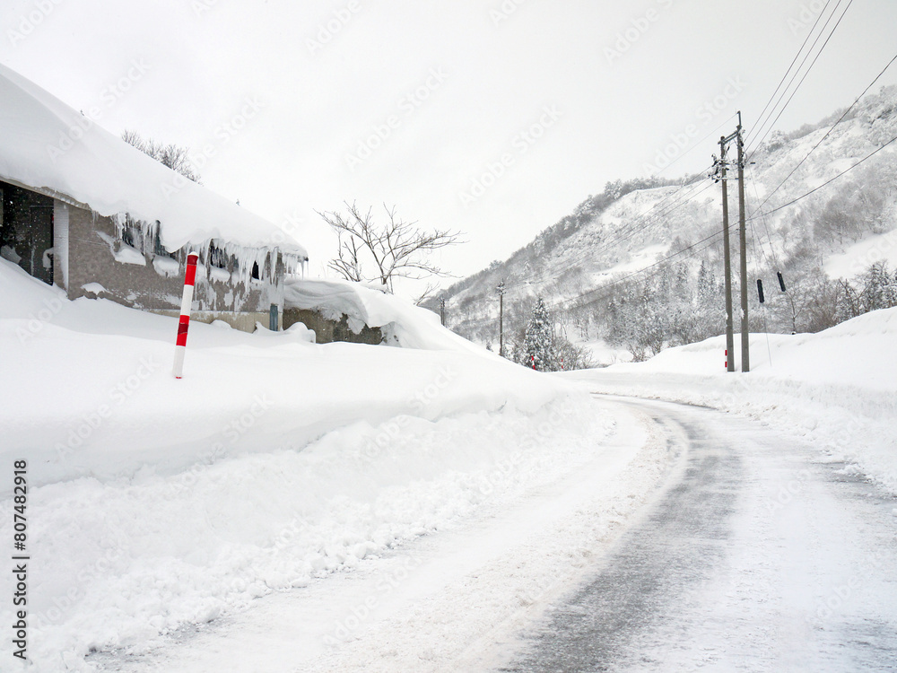 Winter snowy white countryside road, house with big snowdrift on the roof, distant mountains, nature of Yamagata, Japan Winter snowy white countryside road, house with big snowdrift on the roof, distant mountains, nature of Yamagata, Japan