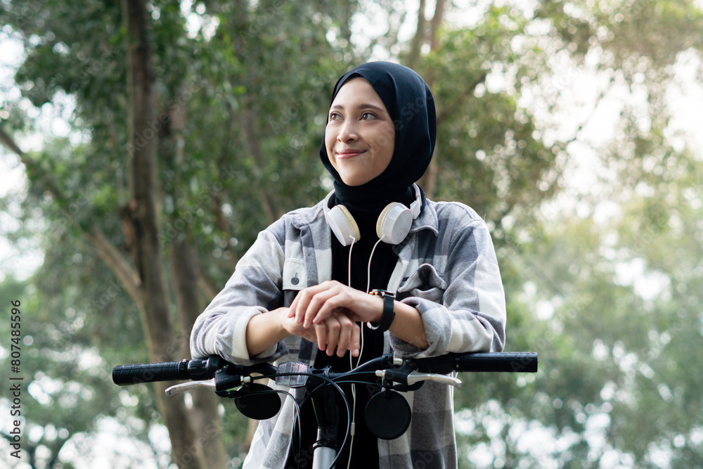Fototapeta premium Young cheerful hijab woman, wearing casual flannel riding bicycle bike on sidewalk in the city park enjoying being outdoors, look aside or ahead. People active urban healthy lifestyle cycling concept.