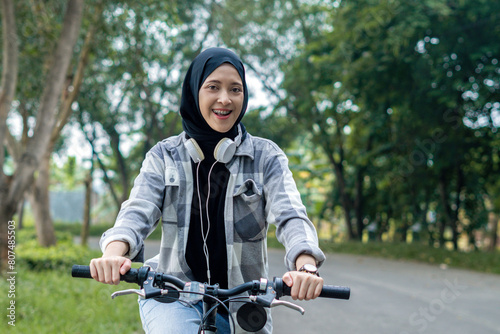 Young cheerful hijab woman, wearing casual flannel riding bicycle bike on sidewalk in the city park enjoying being outdoors, look aside or ahead. People active urban healthy lifestyle cycling concept.