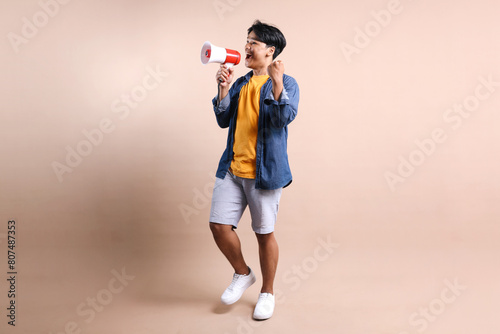Full Length Of Young Asian Man Shouting Through a Megaphone Say Yes Isolated On Beige Background © Queenmoonlite Studio
