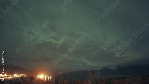 Northen lights in the cloudy sky above a brightly lit town and port in Northern Norway. photo