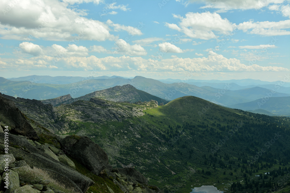Fototapeta premium A look from a rocky peak at a fragment of a picturesque lake sandwiched by high mountains under a cloudy summer sky.