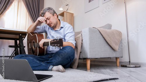 Guitar learning. Educational problem. Virtual lesson. Upset displeased man sitting on floor studying at laptop video course playing chords in home interior copy space. © golubovy