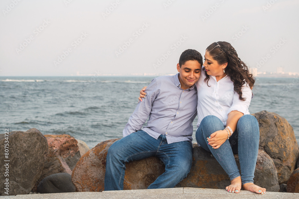 Young Latin woman hugging her teenage son sitting on the rocks in front of the sea during Mother's Day