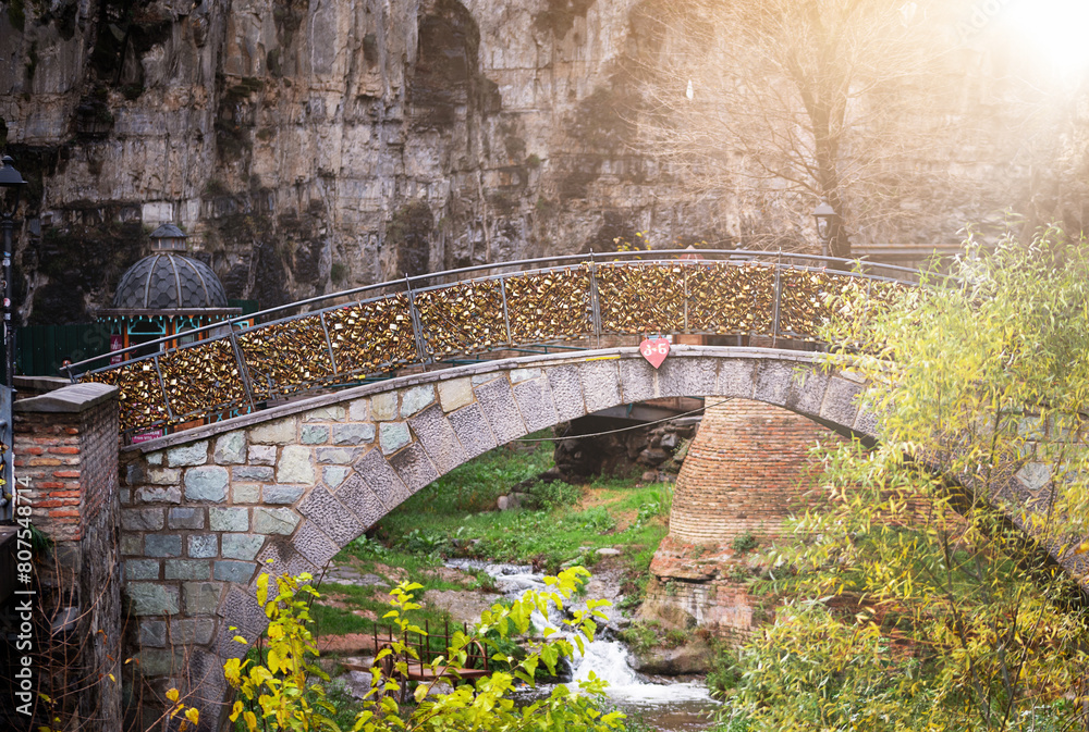 a sunlit morning scene features a charming stone bridge arching gracefully over a placid stream framed by lush foliage against a rugged rock backdrop. Tbilisi Old Town