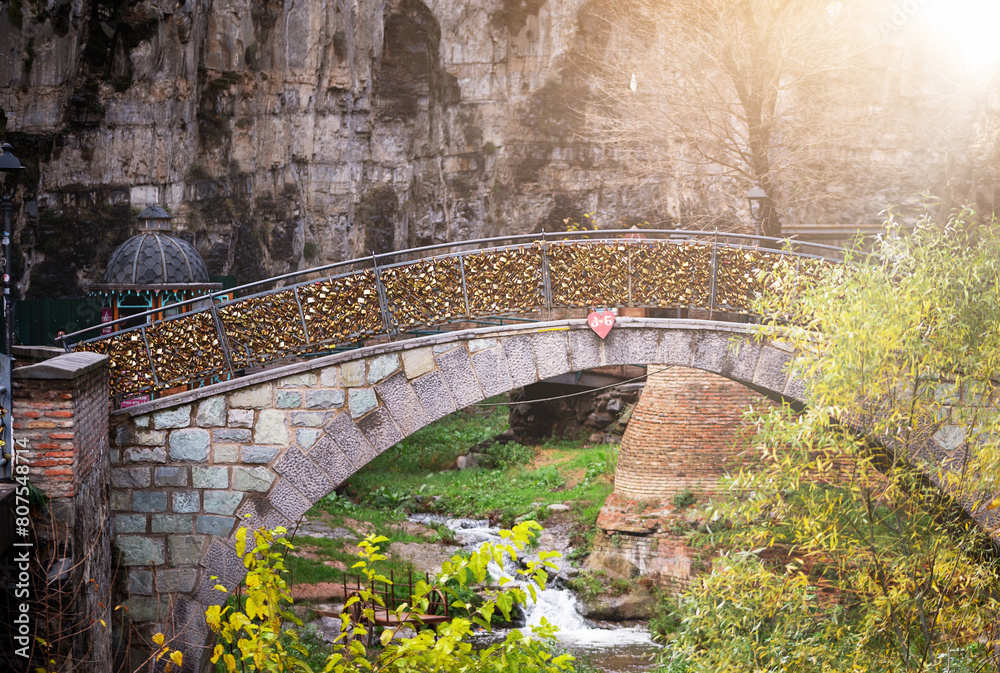 a sunlit morning scene features a charming stone bridge arching gracefully over a placid stream framed by lush foliage against a rugged rock backdrop. Tbilisi Old Town a sunlit morning scene features a charming stone bridge arching gracefully over a placid stream framed by lush foliage against a rugged rock backdrop. Tbilisi Old Town