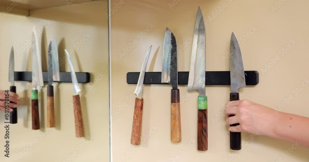 Handcrafted metal knives hang on magnetic holder affixed to kitchen wall. Their varied shapes, including chef's knife and fillet knife. Female hand reaches for each knife in turn, taking and returning Handcrafted metal knives hang on magnetic holder affixed to kitchen wall. Their varied shapes, including chef's knife and fillet knife. Female hand reaches for each knife in turn, taking and returning