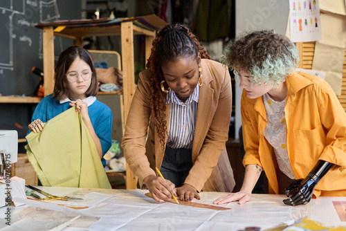 Portrait of Black young woman teaching two girls with disabilities sewing clothing in inclusive atelier studio