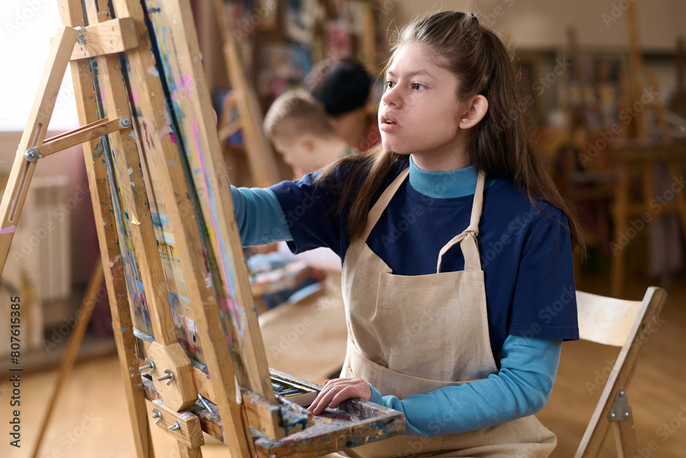 Obraz premium Side view portrait of young girl with disability enjoying art class in studio and painting on canvas