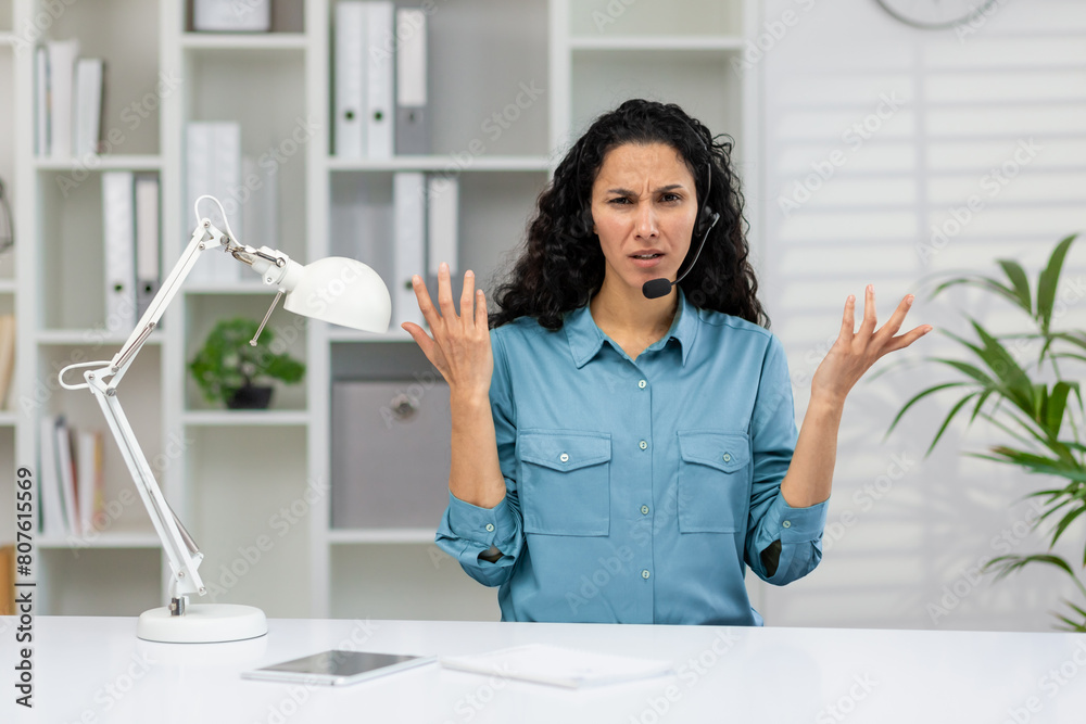 A Hispanic woman looks exasperated while wearing a headset at her office desk, surrounded by modern office decor. A Hispanic woman looks exasperated while wearing a headset at her office desk, surrounded by modern office decor.