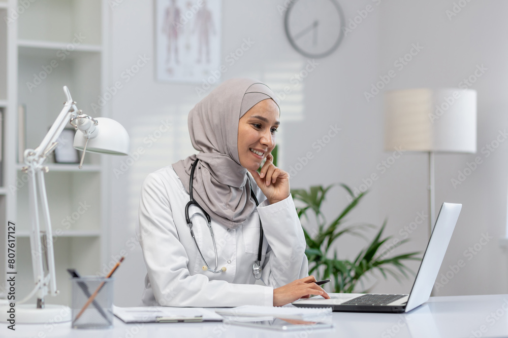 A cheerful Muslim female doctor in hijab engages with a patient online, sitting confidently at her desk in a well-lit modern clinic.