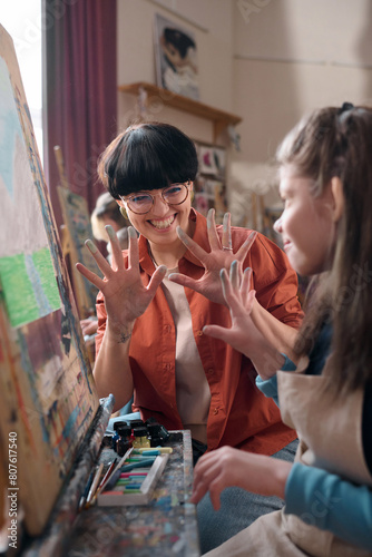 Vertical portrait of playful young woman as female teacher in art class for children showing hands with smudged colors and enjoying fingerpainting photo