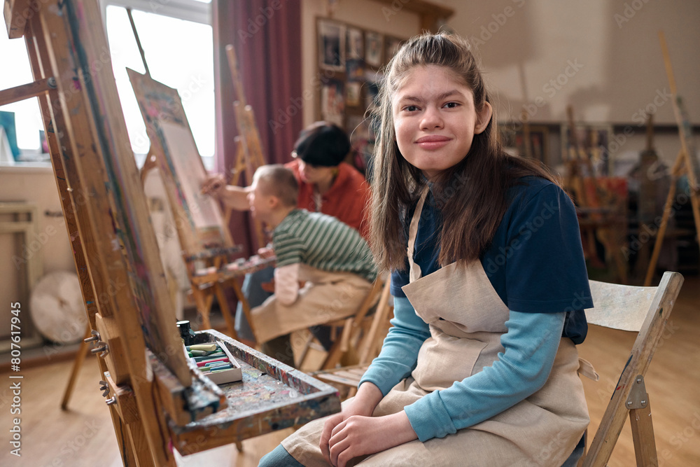 Portrait of teen girl with disability smiling at camera sitting by easel in art class copy space Portrait of teen girl with disability smiling at camera sitting by easel in art class copy space