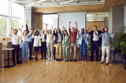 Team of happy people having fun after a business training meeting. Group of excited diverse multiethnic participants standing in a modern office with a projection screen, raising fists and cheering