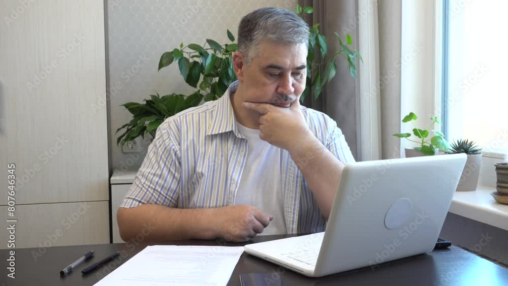 A man works at home on a computer. He looks thoughtfully at the screen, solving the problem. Suitable for content about freelancing or remote work. A man works at home on a computer. He looks thoughtfully at the screen, solving the problem. Suitable for content about freelancing or remote work.
