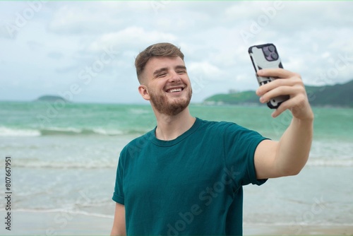 Young guy, handsome happy man at sea is taking picture of hisself, selfie at camera of his phone, using smartphone for social media at summer beach in tropical exotic country. Blogger, vlog photo