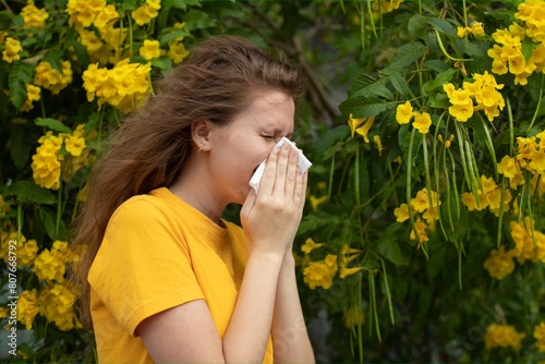 Portrait of beautiful young allergic woman is suffering from pollen allergy or cold on natural flower flowering tree background at spring or sunny summer day sneezes, blowing her runny nose rubs eyes