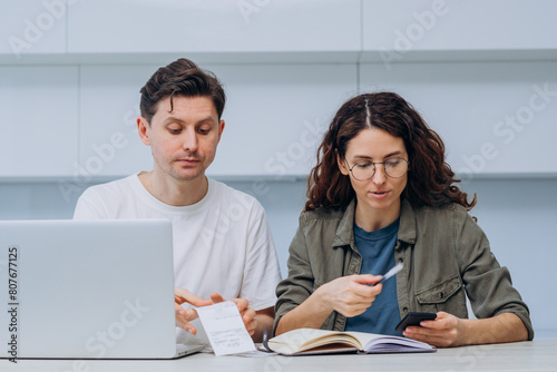 Focused couple sits together at their kitchen table, analyzing financial documents and using a laptop and smartphone to plan their budget.