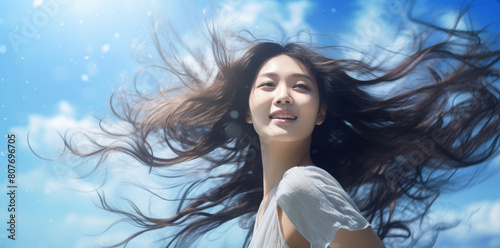 Happy young asian woman smiling with long brown hair blown by the wind on spring summer sunny day on blue sky background. Cheerful and pleased chinese, korean or japanese girl outdoors in nature.