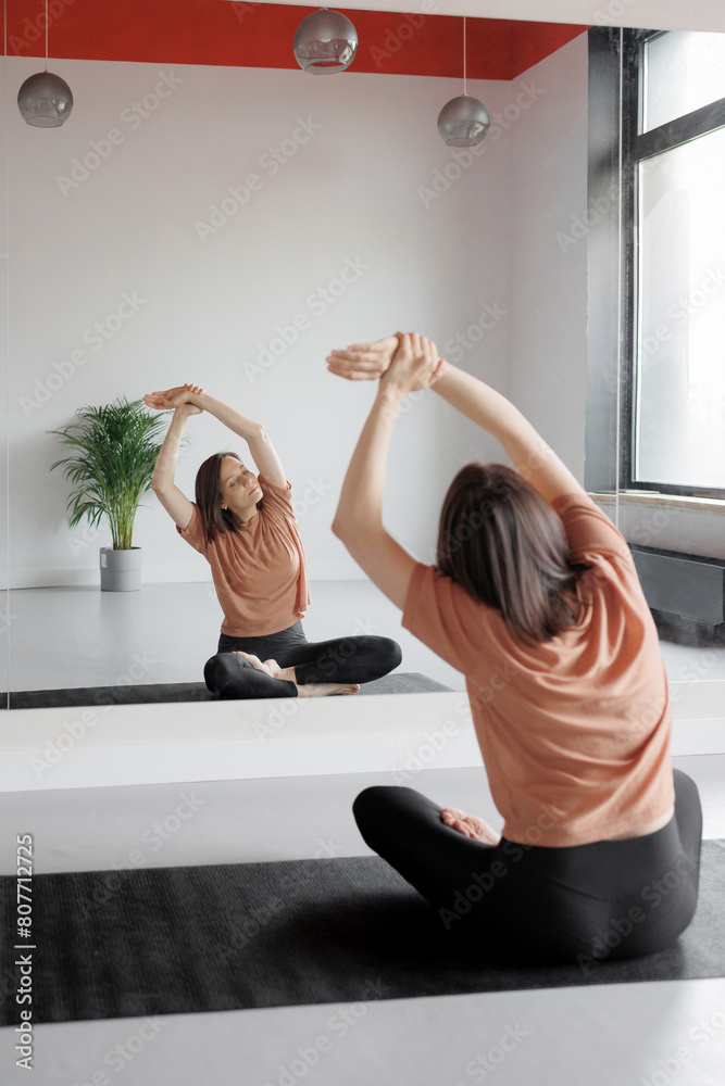 Obraz premium woman in front of mirror does stretching, practices yoga and meditation in gym. Taking care of physical and mental health. European person at sports training session