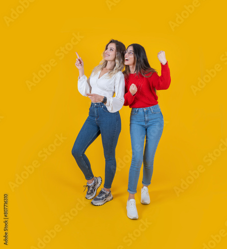 Two excited young full body view caucasian attractive women friends 20s wear casual clothes jeans pointing aside on copy space workspace area mock up do winner gesture isolated yellow background. © Designerant
