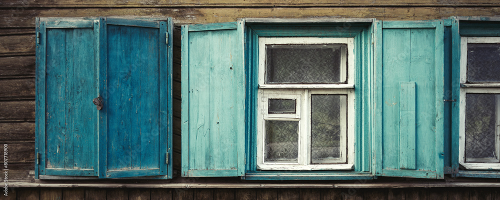 Windows in an old wooden house. Open and closed shutters on the windows. Weathered, faded paint on the wall of the house and on the shutters. Country style. Wide panorama for design and background.