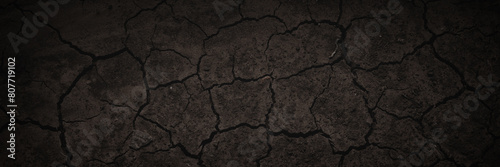 Texture of cracked dried soil. Dark wide panoramic background. Dry ground with cracks. Dark brown rough surface of the soil during summer drought. Shaded texture with vignette. Background for design.