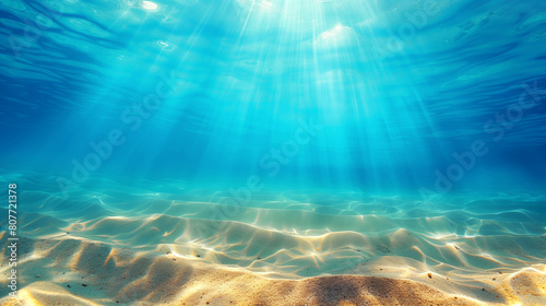 Underwater view of a sunlit sandy sea floor with clear blue water and light rays.