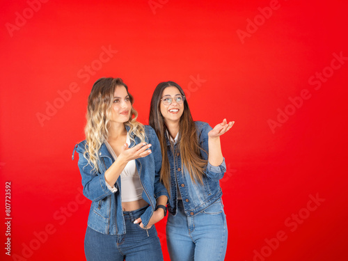 Two young beautiful caucasian women smiling looking to copy space workspace area wear trendy summer jean clothes jackets. Carefree female friends posing over red studio background.