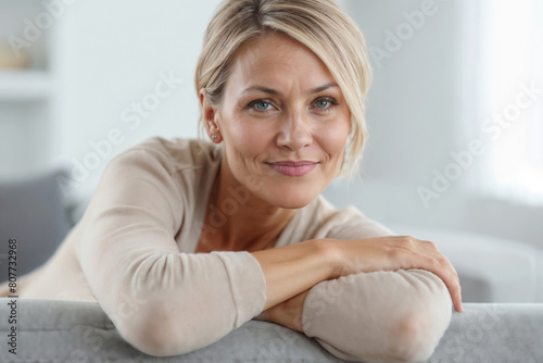 A beautiful 40-year-old woman is relaxing on a light gray sofa against the blurred background of a bright living room