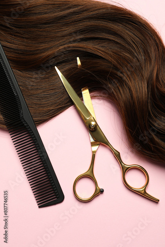 Professional hairdresser scissors and comb with brown hair strand on pink background, top view