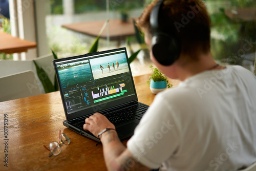 Professional video editor works on laptop with editing software, headphones on. Editing beach scene timeline, focus on creative process in home office. Casual work environment, multimedia project. photo