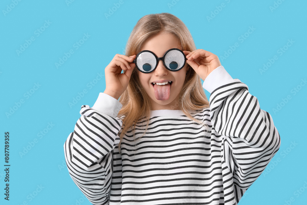 Little girl in funny eyeglasses showing tongue on blue background. April Fools' Day celebration