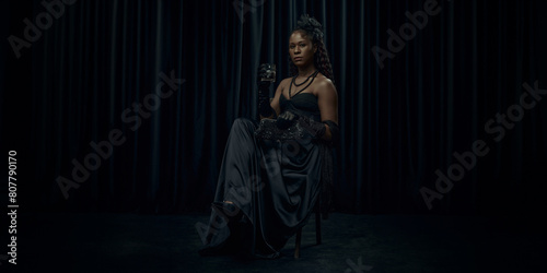 African-American woman, dressed old-fashion dress, looks medieval person, sits confidently on chair holding glass of brandy against dark curtain backdrop. Concept of comparison of eras, vintage. Ad