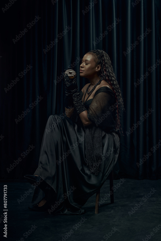 African-American woman in vintage outfit, looks as medieval person holds chocolate donut against dark curtain backdrop. Concept of comparison of eras, modernity and history. Ad
