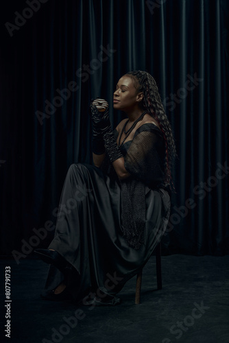 African-American woman in vintage outfit, looks as medieval person holds chocolate donut against dark curtain backdrop. Concept of comparison of eras, modernity and history. Ad © Lustre