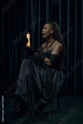 Shocked young African-American woman dressed as medieval person holds modern street food, hot-dog against dark curtain backdrop. Concept of comparison of eras, modernity and history.
